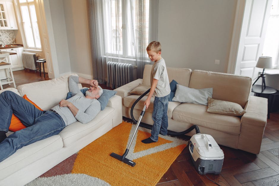 A young boy is vacuuming a tan fabric sofa in a living room using a handheld vacuum cleaner, while an older man with grey hair and glasses is lying on the sofa, resting with his eyes closed and hand on his forehead. The room features wooden flooring partially covered by a colorful rug, large windows with sheer curtains allowing natural light, and neutral-colored walls. The setting displays a clean, well-maintained domestic space with soft pillows on the sofa, and indoor furniture including a side table with a lamp. Carpet Cleaning Paddington services are highlighted by the emphasis on surface cleaning and maintaining hygiene in a residential setting.
