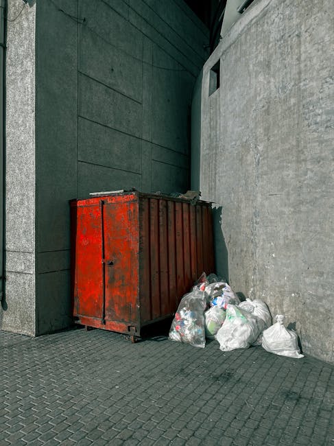 An outdoor scene in the Paddington area showing a red metal waste container positioned against a gray textured wall with a darker gray, paneled surface on the left side. The container appears weathere