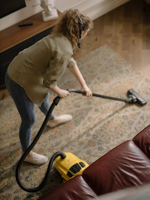 A person wearing a full white protective suit, gloves, and a face mask is using a vacuum or steam cleaning machine on a beige carpet in a well-lit living room. The room features light-colored walls, a