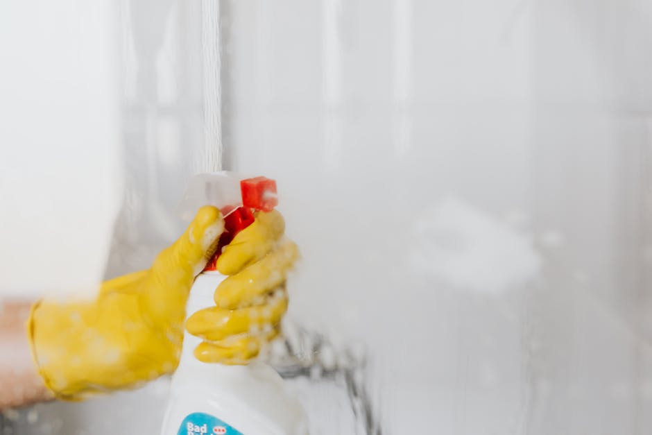 Close-up of a hand wearing a yellow rubber glove holding a spray bottle of cleaning solution, aimed at a tiled wall in a kitchen or bathroom. The spray bottle is partially obscured by foam and mist from the spray, indicating surface cleaning activity. The tiled wall is made of light-colored ceramic tiles, appearing clean and shiny, with some wet streaks visible. The lighting is natural and bright, highlighting the cleanliness of the surface. The scene suggests a thorough sanitisation process typical of domestic or commercial cleaning practices, as performed by Carpet Cleaning Paddington, especially in the context of surface and deep cleaning of tiled surfaces.