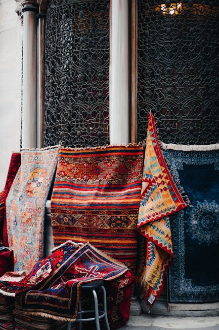 A collection of traditional woven rugs and carpets with intricate geometric and floral patterns, displayed outdoors against a decorative black metal fence with ornate scrollwork. The rugs feature vibrant colors including reds, blues, oranges, and cream, and are arranged overlapping one another, with some draped over a metal chair or leaning against the fence. The setting appears to be in a courtyard or outdoor market area, with natural diffused lighting highlighting the detailed textures and vivid hues of the textiles, which are being prepared or displayed for cleaning or restoration by Carpet Cleaning Paddington, focusing on surface cleaning and preservation of fabric materials.