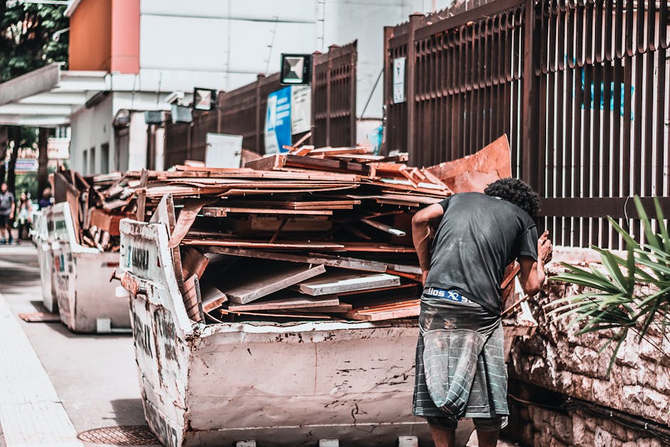 A man wearing a dark grey T-shirt and plaid shorts stands outdoors beside a large white skip filled with broken tiles, wooden planks, and construction debris. The skip is positioned on a city sidewalk