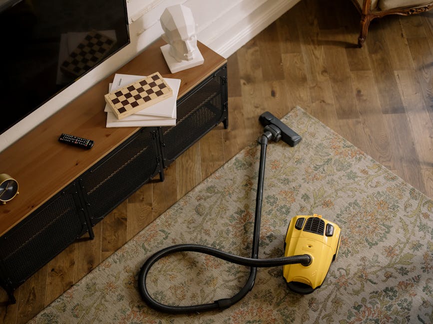 An interior scene showing a vacuum cleaner on a patterned area rug in a living room with wooden flooring. To the left, a black wooden sideboard with a small statue, chessboard puzzle, papers, and a remote control on top. The room is well-lit, highlighting the clean, polished surfaces including the wooden floor and the rug, which appears free of dust and debris. The image emphasizes surface cleaning and maintenance, aligned with domestic cleaning services by Carpet Cleaning Paddington in the W2 area, focused on deep cleaning and sanitisation practices.