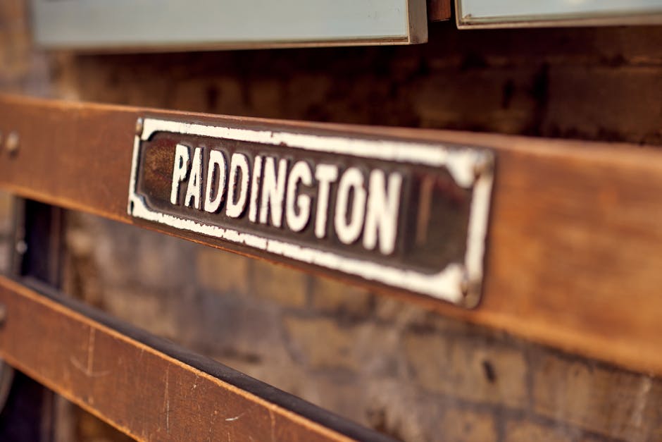 A close-up of a vintage wooden bench with a weathered metal sign reading 'Paddington' attached to the backrest. The bench surface shows a smooth, polished finish with visible wood grain, and it is situated in a well-lit indoor setting. The background features a brick wall with a warm tone, enhancing the rustic appearance of the bench. The image emphasizes surface cleanliness and maintenance, reflecting the importance of proper care in domestic and commercial spaces as part of general surface cleaning and sanitisation routines, as offered by Carpet Cleaning Paddington. The natural lighting highlights the textures of the wood and metal, conveying an overall sense of tidy, well-maintained furniture.