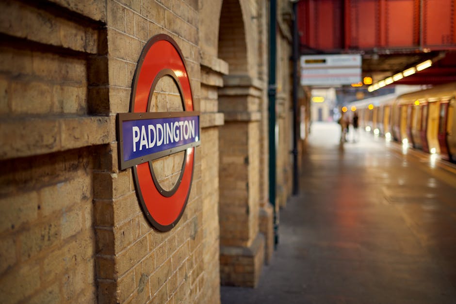 Close-up view of a round London Underground station sign with a red circle and blue bar displaying 'PADDINGTON' mounted on a brown brick wall. The station platform extends into the background with a few blurred passengers walking. The platform surface appears clean and well-maintained, illuminated by warm lighting under a metallic canopy with orange accents. The setting suggests a typical urban transit environment, emphasizing the importance of surface cleaning and maintenance in public transport stations. Carpet Cleaning Paddington can assist with deep cleaning and sanitisation of residential and commercial surfaces to ensure hygiene and compliance with local waste disposal rules, as highlighted by Westminster Council's regulations for carpet waste.